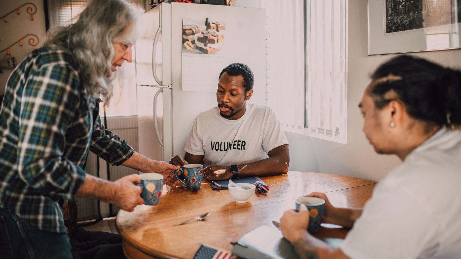 A man in a volunteer T-shirt shares coffee with two people at a kitchen table